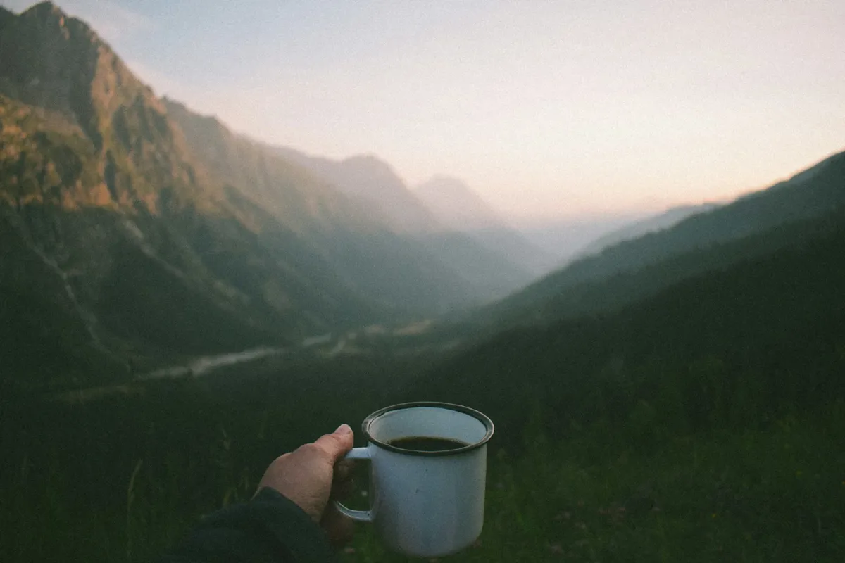 Hand hält eine Emaille-Tasse mit Kaffee vor einem Bergtal im weichen Morgenlicht