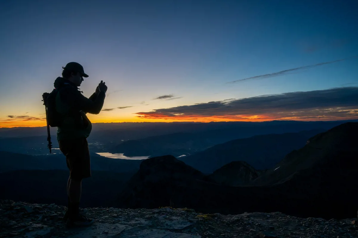 Silhouette eines Wanderers mit Rucksack, der auf einem Gipfel den farbigen Horizont über Tälern und einem See fotografiert