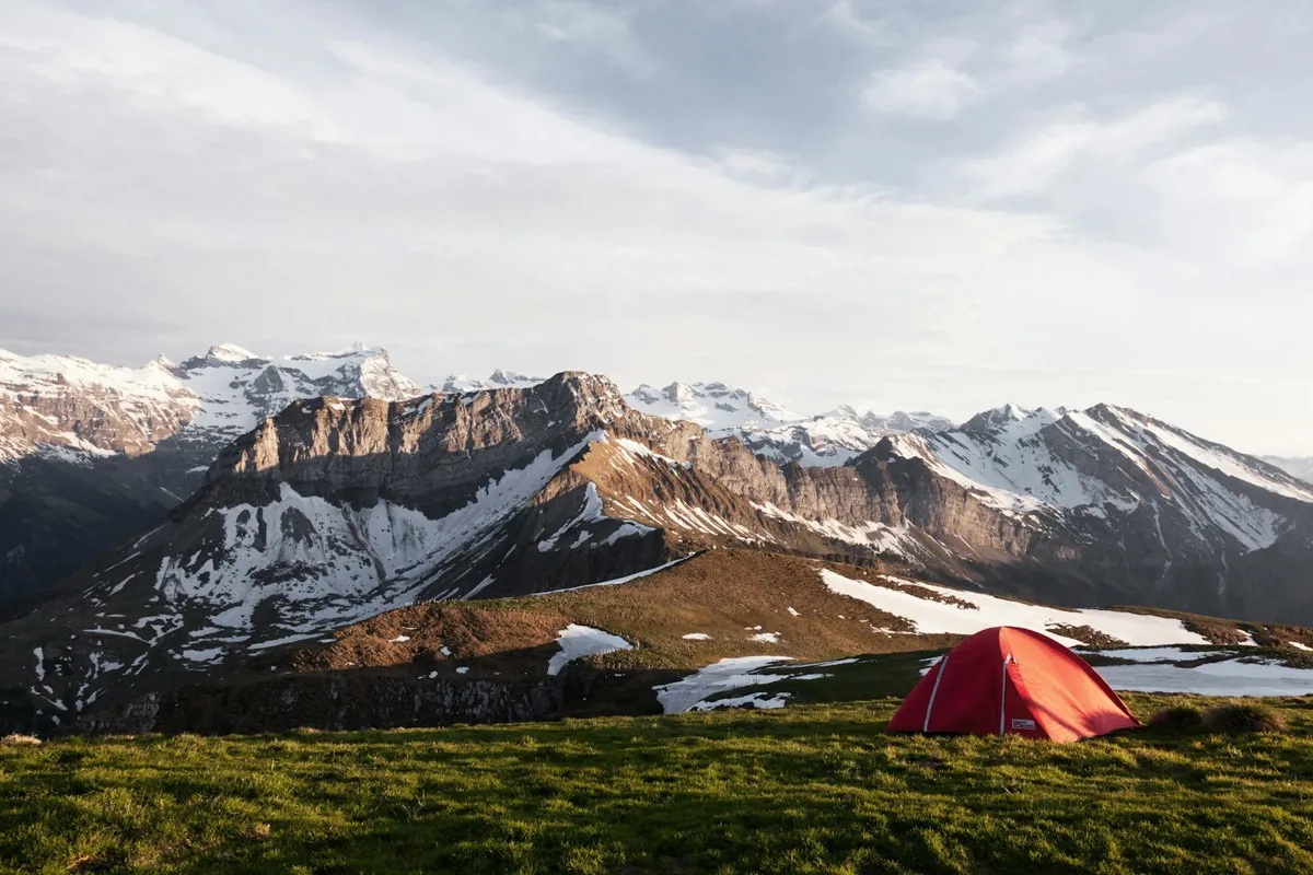 Rotes Zelt auf einer Wiese vor einer Bergkette mit Schneefeldern unter bewölktem Himmel