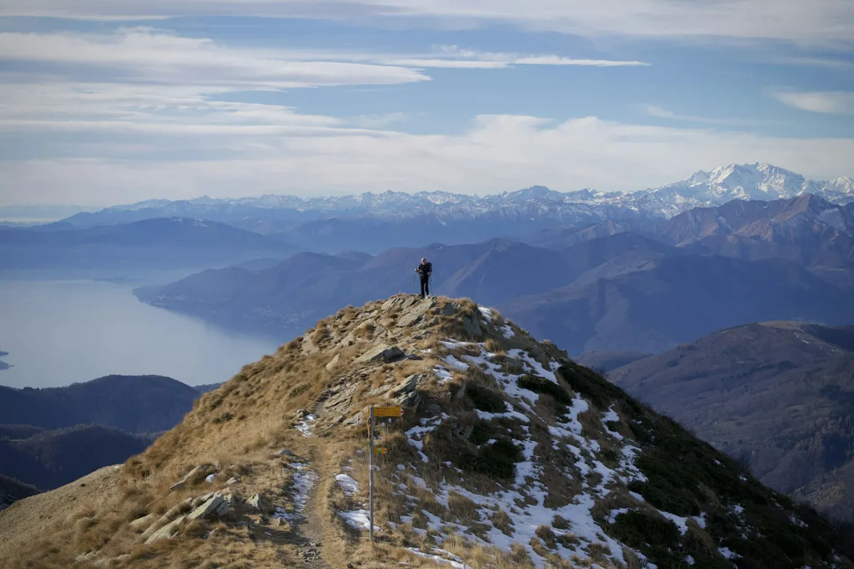 Wanderer steht auf einem grasigen Gipfelgrat mit Schneeresten, dahinter ein See und weite Bergketten bis zu verschneiten Gipfeln