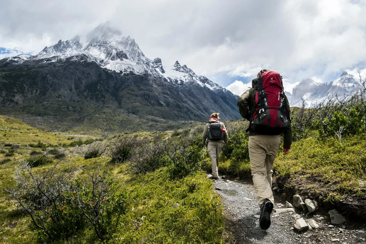 Zwei Wanderer mit Rucksäcken gehen von hinten gesehen einen Schotterpfad entlang, vor ihnen wolkenverhangene, verschneite Gipfel